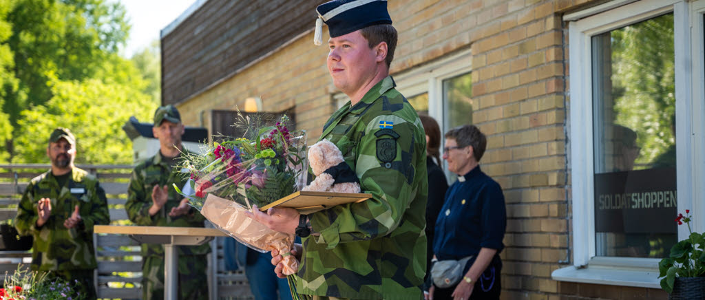 Soldat står i förgrunden med blommor och en nalle. I bakgrunden står två militärer och klappar händerna samt en civilklädd kvinna. 